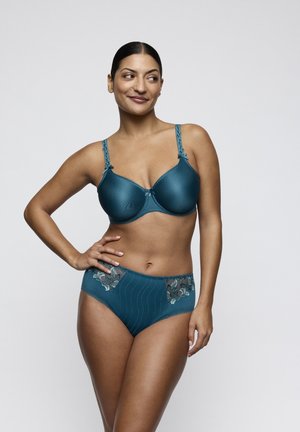 Vrouw staat met hand op heup, draagt turquoise beha met kanten bandjes en bijpassend hooggetailleerd ondergoed met bloemrijke borduring op lichte achtergrond.