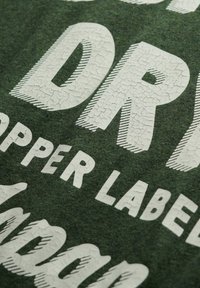 Grønne stof med store, revnede hvide bogstaver der læser "DRY", med yderligere tekst i mindre skrifttype. Glat tekstur.
