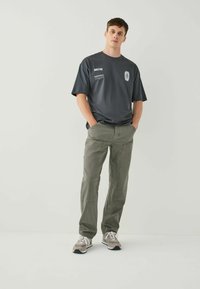 Gray cotton t-shirt with text graphics, paired with light olive cargo pants. Model wears gray and white sneakers. Simple design, relaxed fit.