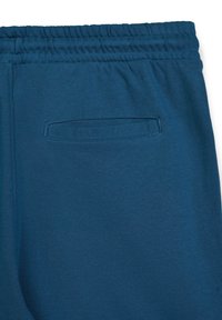 Des joggeurs en coton bleu présentent une taille élastique et une poche arrière unique avec un rabat. Texture lisse avec une couleur unie et homogène.