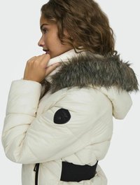 Pufová bunda v off-white farbe s kožušinovou kapucňou. Má čiernu logo nálepku na ramene a elastický pás pre bezpečné uchytenie.