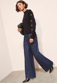 Pulover negru cu m�âneci lungi și detalii cu nasturi pe mâneci, asortat cu blugi denim albaştri cu picioare largi și pantofi cu vârf ascuțit negri.