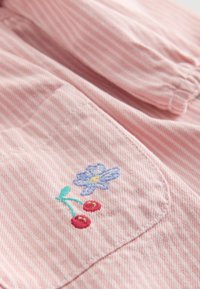 Różowa tkanina w paski z haftowanym kwiatem w odcieniach niebieskiego i zielonego, w towarzystwie czerwonych wiśni. Posiada kieszeń z widocznym ściegiem.