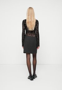 Top preto transparente de mangas longas com padrão de flores, combinado com uma saia texturizada cinza. Modelo usa sapatos pretos e collants. Vista traseira.