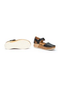 Zapatos de cuero negro con diseño trenzado, correa ajustable, entresuela de corcho y suela de goma blanca con marca en relieve.