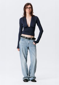 Marineblaues Wickeloberteil mit langen Ärmeln, kombiniert mit hellblauen, weit geschnittenen Jeans und schwarzem Gürtel. Das Modell trägt eine Brille und steht vor einem neutralen Hintergrund.