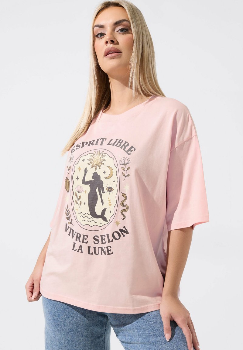 Femme blonde portant un T-shirt rose ample avec un dessin de sirène, soleil, lune, plantes, et texte français "Esprit Libre Vivre Selon La Lune".