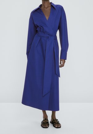 Woman wearing a long-sleeve, deep blue wrap dress with a tied waist belt and black double-buckle flat sandals, standing on a plain background.