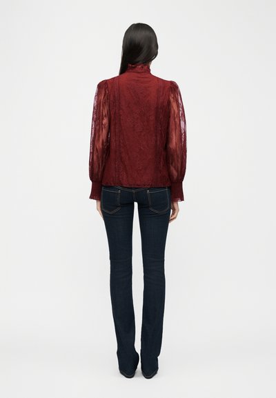 Γυναίκα που φοράει διάφανη μπορντό μπλούζα από δαντέλα με μακριά μανίκια φουσκωτά και σκουρό μπλε εφαρμοστά τζιν, στέκεται με την πλάτη προς το μέρος μας σε λευκό φόντο.