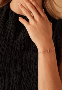 Zilveren armband met twee in elkaar grijpende hartvormen en een delicate ketting, gedragen om een pols tegen een gestructureerde zwarte stof.