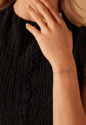 Zilveren armband met twee in elkaar grijpende hartvormen en een delicate ketting, gedragen om een pols tegen een gestructureerde zwarte stof.