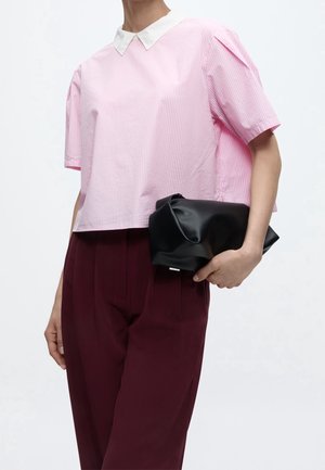 Persona con una blusa rosa de manga corta con cuello blanco y pantalones burdeos, sosteniendo un bolso clutch negro suave en una mano.