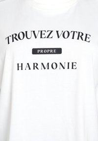 White t-shirt featuring black printed text that reads "TROUVEZ VOTRE PROPRE HARMONIE." Round neckline and soft cotton fabric.