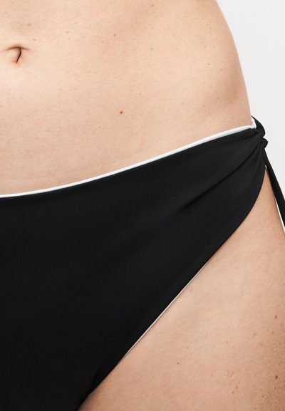 Κοντινό πλάνο στη μέση ενός ατόμου που φορά μαύρο κάτω μέρος μπικίνι με λευκή λεπτομέρεια περιγράμματος και δεσίματα στο πλευρό, με ουδέτερο φόντο.