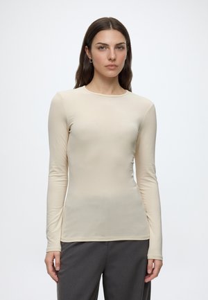 Vrouw met lang bruin haar, gekleed in een aansluitend beige shirt met lange mouwen en donkergrijze broek, staand tegen een effen witte achtergrond.