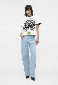 Tânără cu păr negru poartă tricou alb cu imprimeu grafic, blugi largi de culoare albastru deschis și pantofi negri cu toc ascuțit, stând în picioare pe fundal alb simplu.