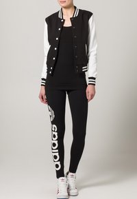Černo-bílá univerzitní bunda s proužkovanými manžetami, přiléhavé černé tílko, černé legíny s bílým logem adidas a bílé tenisky.