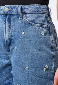 Vaqueros de mezclilla azul con bordado floral, con margaritas y hojas verdes. Diseño clásico de cinco bolsillos, costuras detalladas y un botón de metal.