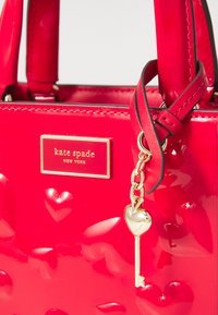 Rode lakleren handtas met een gouden logo plaat, hartvormige accenten en een sleutelbedeltje dat met een delicate ketting is bevestigd.