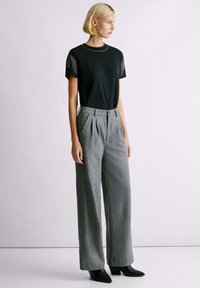Camisa negra de manga corta con detalle de flecos en los hombros, combinada con pantalones grises de pierna ancha y pliegues, y botas de tacón alto negras.