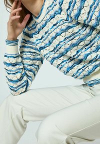 Sweter dzianinowy w kremowym kolorze z niebieskimi falistymi wzorami. Posiada teksturowany materiał i ściągacze z kolorowymi akcentami. Zestawiony z białymi spodniami.