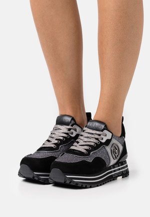 Jambes portant des baskets compensées noires et argent avec des semelles texturées et des lacets métalliques argentés sur un fond uni.