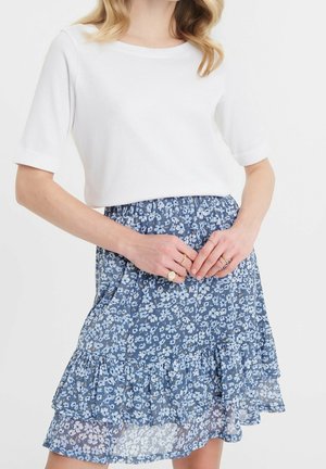 Kvinde iført en hvid top med korte ærmer og et lagdelt blåt blomsterprint nederdel, med hænderne foldet og flere ringe på fingrene.