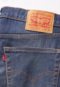 Jean en denim bleu avec une étiquette en cuir marron comportant un texte imprimé rouge. Coupe slim, design à jambes droites, poches standard et couture distinctive.
