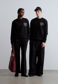 Las sudaderas negras tienen un pequeño gráfico en el pecho. Ambos llevan pantalones negros holgados. La mujer lleva un bolso acolchado de color burdeos.