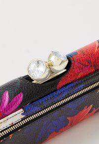 Nahkalaukku kukkakuviolla, jossa on kultainen vetoketju ja kaksi kirkasta kristallikoristetta ylhäällä. Vallitsevat värit ovat punainen, sininen ja musta.