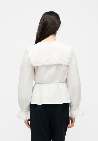 Femme aux longs cheveux foncés portant un chemisier blanc avec un col et des poignets de marin ornés de dentelle, assorti à un pantalon foncé, vue de dos.