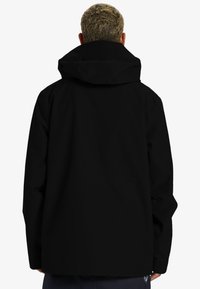 Chaqueta negra con capucha y textura suave, presenta un corte holgado y mangas largas. La parte trasera tiene un diseño minimalista sin logotipos visibles.