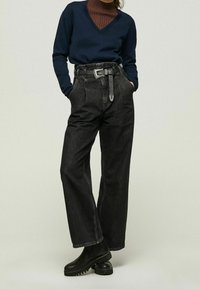 Schwarze hoch taillierte Jeanshose mit weitem Bein, ausgestattet mit einem geschnürten Gürtel und eingenähten Akzenten. Kombiniert mit einem marineblauen V-Ausschnitt-Pullover.