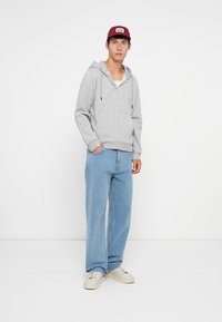 Grauer Reißverschluss-Hoodie mit Kordelzügen, Fronttaschen, kombiniert mit hellblauen weiten Jeans und weißen Sneakers, vor einem einfarbigen Hintergrund.