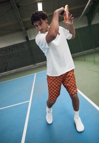 Tenisový hráč v bielom tričku a oranžových kockovaných šortkách, držiaci tenisovú raketu, na modrom kurte s zeleným pozadím.