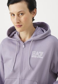 Lila zip-uppi kapuca narejena iz mehkega blaga, ki ima kenguru žep in izšito EA7 logo na prsih. Vključuje kapuco z vrvico.