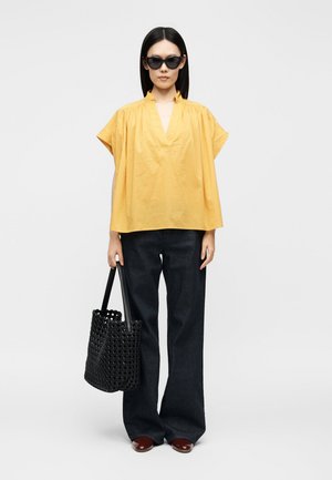 Mujer con blusa amarilla oversize, vaqueros oscuros de pierna ancha, gafas de sol, zapatos marrones y sosteniendo un bolso negro tejido sobre fondo blanco.