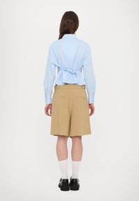 Helles blaues Hemd mit Rückenschnürdetail, kombiniert mit beigen Shorts mit seitlichen Taschen und glatter Textur, ergänzt durch schwarze Schuhe.