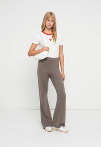Chemise blanche à manches courtes avec un col rouge et un logo, associée à un pantalon ample marron. Le modèle tient un sac à épaule blanc.