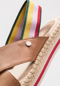 Sandale en cuir marron avec des bretelles en tissu rayé multicolore, semelle en jute, et un bouton logo décoratif. Présente une forme arrondie et des accents contrastés.