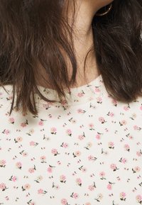 Top a fantasia floreale color crema con piccoli fiori rosa e blu, realizzato in tessuto morbido. Presenta un collo rotondo e maniche corte.