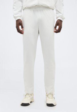 Άνδρας που φοράει λευκό παντελόνι jogging με ελαστική μέση και εκρού αθλητικά παπούτσια, στέκεται με χαλαρά τα χέρια στο πλάι, σε λευκό φόντο.