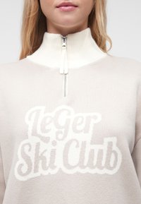 Bézs színű félcipzáras pulóver fehér bordázott gallérral. A mellkason világos bézs színű, strukturált "LeGer Ski Club" felirat található.