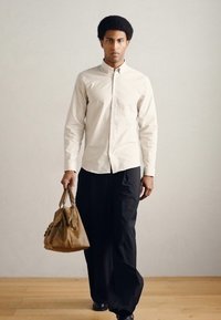 Μπεζ πουκάμισο με κουμπιά, μαύρο παντελόνι φαρδύ, καφέ δερμάτινη τσάντα με μεταλλικές λεπτομέρειες, σε μοντέλο που περπατά σε ξύλινο πάτωμα μπροστά σε ένα ουδέτερο τοίχο.