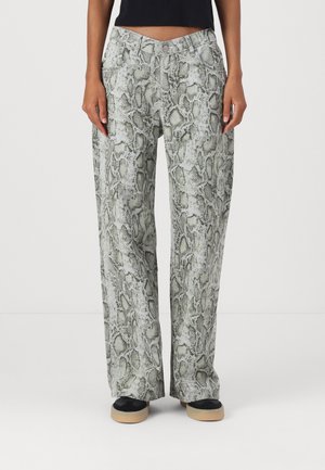Femme portant un pantalon taille haute à jambes larges avec un motif peau de serpent gris et noir, associé à un haut noir et des chaussures noires avec des semelles beiges.