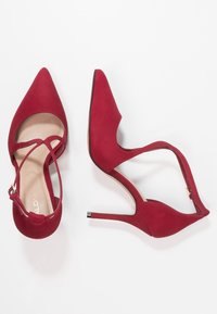 Ett par röda högklackade kvinnor skor med spetsiga tår och ankelsnören på en vit bakgrund.