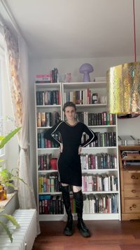 Abito nero a maniche lunghe con strisce bianche lungo i fianchi, abbinato a stivali neri al ginocchio. Sullo sfondo ci sono scaffali di libri e decorazioni.