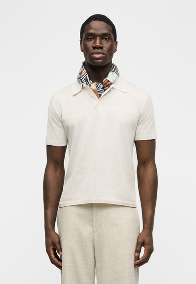 Άνδρας που φοράει μπεζ κοντομάνικη μπλούζα πόλο, ασορτί ανοιχτό μπεζ παντελόνι και ένα πολύχρωμο φουλάρι με περίτεχνο μοτίβο γύρω από το λαιμό του, στέκεται μπροστά σε λευκό φόντο.