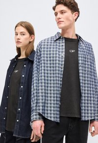 Νέος άνδρας και γυναίκα φορούν μαύρα πουκάμισα κάτω από ανοιχτά casual ρούχα, στέκονται κοντά μπροστά σε ένα απλό φωτεινό φόντο.