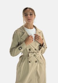 Manteau trench beige à double boutonnage avec de grands revers, une ceinture à la taille et des détails de boutons. Tissu lisse avec une coupe structurée.